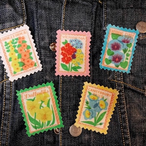 May include: Five floral-themed stamp-shaped pins with scalloped edges are displayed on a denim jacket. The pins feature images of flowers like hollyhocks, verbena, and daffodils, with text labels. The color palette includes pink, green, yellow, and blue.