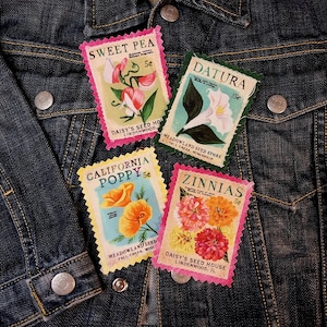 May include: Four vintage-style seed packet patches on a dark denim jacket. The patches feature floral illustrations of Sweet Pea, Datura, California Poppy, and Zinnias. Each patch has a different color border and text indicating the flower name and price.
