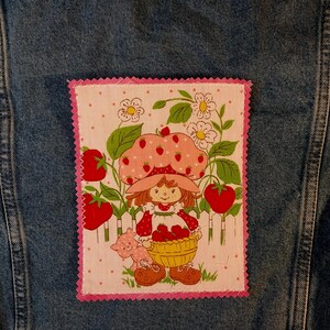 May include: A rectangular fabric patch with pink zigzag edges featuring a cartoon character wearing a strawberry-patterned hat, holding a basket of strawberries, and standing near a white fence, all set against a light pink polka dot background.