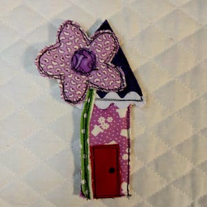 May include: A fabric applique shaped like a house with a purple floral pattern, a red door, and a green side panel. A large, five-petaled light purple flower with a dark purple center is attached to the top, featuring black stitched outlines.