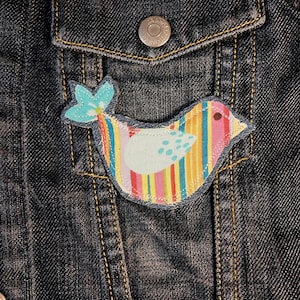 May include: A fabric bird-shaped patch featuring vertical stripes in pink, yellow, blue, and green, with a light blue wing decorated with teal dots. The patch is stitched onto a dark blue denim jacket with a visible metal button on the pocket flap.