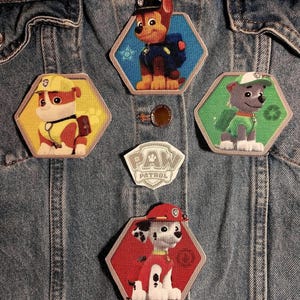 May include: A collection of five embroidered patches featuring Paw Patrol characters. Four hexagonal patches display cartoon dogs in yellow, blue, green, and red, while a central shield-shaped patch features the white Paw Patrol logo text.