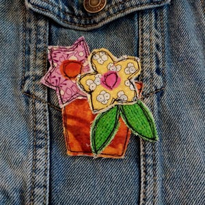 May include: A blue denim jacket featuring a fabric appliqué of a potted plant. The appliqué includes a brown pot, two green leaves, a yellow flower with white floral patterns, and a pink flower with a dark center, all with visible black stitching.