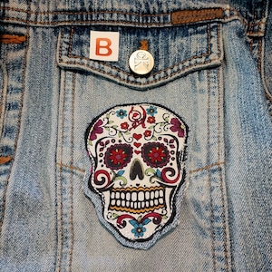 May include: Light blue denim jacket with a colorful Day of the Dead skull patch. The patch features a white skull with floral and heart designs in red, purple, and blue. A small "B" sticker and a silver button are on the pocket.