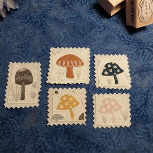 May include: Five square fabric patches with pinked edges, each featuring a different mushroom illustration in gray, orange, dark blue, yellow, or pink. Two wooden rubber stamps are positioned nearby, one with wavy lines and one labeled PAR AVION.