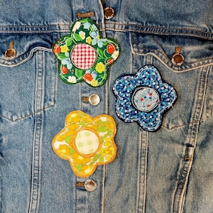 May include: Three fabric flower patches attached to a blue denim jacket. The top left patch is green with red strawberries and a red gingham center. The right patch is blue with small floral patterns. The bottom patch is yellow with a green gingham center.