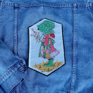 May include: A blue denim jacket featuring a large, rectangular fabric patch sewn onto the back. The patch displays a colorful illustration of a figure in a green bonnet and boots holding flowers, with the text Holly Hobbie printed at the bottom.
