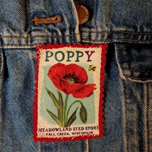 May include: A denim jacket with a Poppy patch. The patch features a red poppy flower illustration with the word "POPPY" in purple and the text "MEADOWLAND SEED STORE FALL CREEK, WISCONSIN". The patch has a red border and a light blue background.
