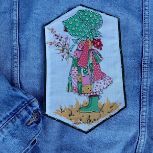 May include: A denim jacket with a colorful patch featuring a girl in a patchwork dress, green boots, and a bonnet, holding flowers. The patch has a black border and the words "Holly Hobbie".