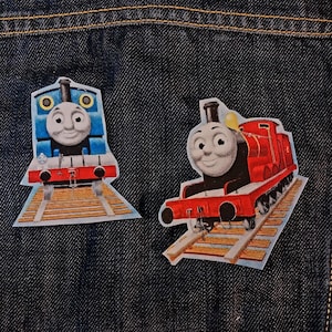 May include: Two iron-on patches featuring Thomas the Tank Engine and James the Red Engine. The blue Thomas engine faces forward, while the red James engine is angled. Both locomotives are depicted on wooden train tracks against a dark denim fabric.