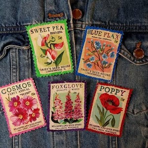 May include: Five vintage-style seed packet patches featuring floral illustrations. The patches include "Sweet Pea," "Blue Flax," "Cosmos," "Foxglove," and "Poppy." Each patch has a unique color scheme and text, with prices indicated.