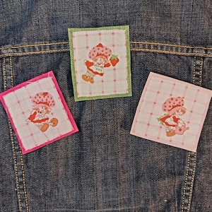 May include: Three square fabric patches featuring a cartoon character with a strawberry hat and dress. The patches are on a denim jacket. One patch has a pink border, one has a green border, and one has a pink border.