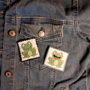 May include: Two square patches with a cartoon frog design are affixed to a denim jacket. The frogs are green with white bellies and red mouths, set against a white background with a decorative border. The jacket is a medium wash blue.