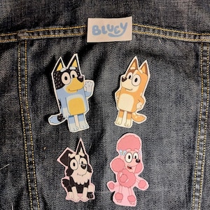 May include: Four cartoon dog stickers featuring Bluey, Bingo, Mackenzie, and Coco, arranged on dark blue denim fabric. A small white rectangular label with the blue text Bluey is positioned above the stickers. Each sticker has a white border.