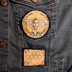 May include: Close-up of a denim jacket with two embroidered patches. The larger patch features a detailed bee illustration on a yellow background, surrounded by leaves. The smaller patch displays a honeycomb design.
