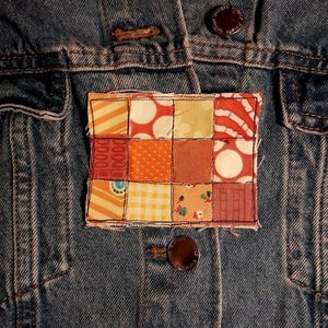 May include: A blue denim jacket featuring a rectangular patchwork applique sewn onto the front. The patch consists of twelve smaller fabric squares in various shades of orange, yellow, and red, featuring patterns like polka dots, stripes, and florals.