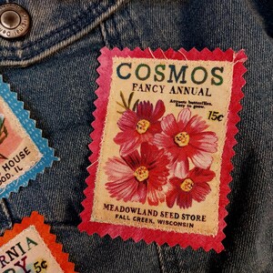 May include: Close-up of a denim jacket adorned with several vintage seed packet patches. The central patch features a pink border and the text "COSMOS FANCY ANNUAL" with a floral illustration and the text "MEADOWLAND SEED STORE".