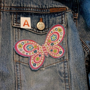 May include: A blue denim jacket featuring a butterfly patch with pink, white, and green floral patterns. A small white square with the letter A is placed near a silver button embossed with the letters CK. The jacket has a brown leather label.