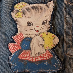 May include: A fabric patch featuring a vintage-style illustration of a kitten wearing a yellow bow and a blue dress with a red and white checkered apron, holding a yellow plate and a white cloth, stitched onto a blue denim fabric background.