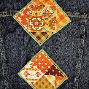 May include: Two diamond-shaped fabric patches sewn onto a dark blue denim jacket. The top patch features a central square with a bird and floral motif surrounded by orange and brown plaid and floral fabric. The bottom patch is a patchwork of floral and geometric patterns in orange, yellow, brown, and white.
