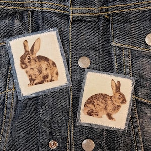 May include: Two square fabric patches with a denim border, each featuring a brown rabbit illustration. The patches are affixed to a dark blue denim jacket. The rabbit design has a vintage, rustic aesthetic.