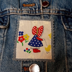 May include: A blue denim jacket featuring a rectangular fabric patch with a white background and a graphic print of a person in a red polka-dot bonnet and blue polka-dot dress walking a yellow duckling, surrounded by colorful flowers.