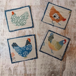 May include: Four square fabric coasters with a denim blue border and stitched edges. Each coaster features a different colorful chicken design. The chickens have various patterns and colors, including blue, orange, and green.