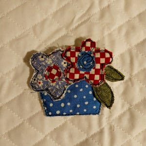 May include: A fabric applique featuring a blue pot with white polka dots containing two flowers. One flower has a light blue floral pattern with a red and white checkered center, and the other has a red and white checkered pattern with a blue center.