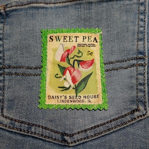 May include: Close-up of a light blue denim jeans pocket with a rectangular patch. The patch features a floral design of sweet peas with the text "Sweet Pea" and "Daisy's Seed House, Lindenwood, IL". The patch has a green, zig-zag border.