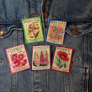 May include: Five vintage-style seed packet patches with floral illustrations and text, including "Sweet Pea," "Blue Flax," "Cosmos," "Foxglove," and "Poppy." The patches are displayed on a denim jacket.