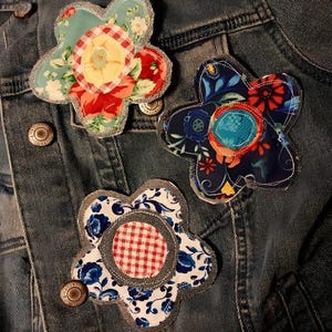 May include: Three fabric flower patches attached to a blue denim jacket. The top left patch features teal, red, and yellow floral patterns with a red gingham center. The right patch has dark blue, red, and orange floral prints. The bottom patch is white with blue flowers and a red gingham center.