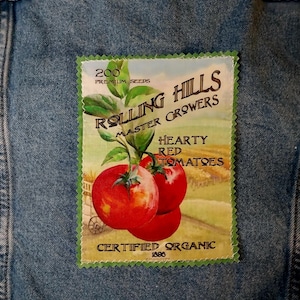 May include: A denim jacket with a rectangular patch featuring a vintage illustration. The patch displays the text "Rolling Hills Master Growers" and "Hearty Red Tomatoes" with images of ripe tomatoes. The patch also includes the text "Certified Organic 1896".