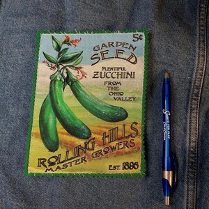 May include: A rectangular fabric patch with a green zigzag stitched border featuring a vintage-style illustration of two green zucchinis. The text reads Garden Seed, Plentiful Zucchini, From the Ohio Valley, Rolling Hills Master Growers, Est. 1896, and 5 cents. A blue ballpoint pen is placed next to the patch for scale.