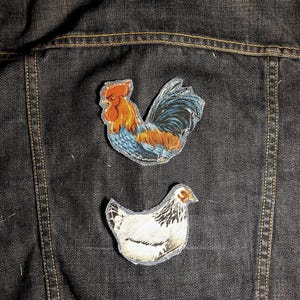 May include: Two fabric patches attached to a dark denim jacket. The top patch features a rooster with orange, red, and blue plumage. The bottom patch depicts a white hen with black neck and tail feathers, showcasing detailed embroidery.