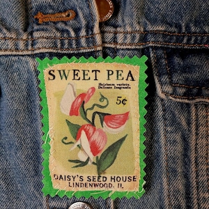 May include: A denim jacket with a Sweet Pea patch. The patch features a floral illustration with red and white flowers, green leaves, and the text "SWEET PEA" and "DAISY'S SEED HOUSE LINDENWOOD, IL."