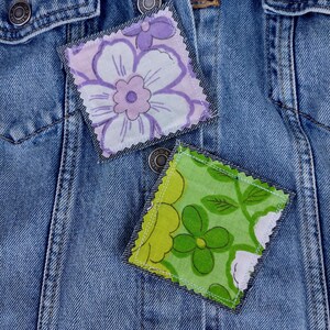 May include: Close-up of a blue denim jacket with two decorative fabric patches. One patch features a purple and white floral design, and the other has a green and white floral pattern. The patches are square-shaped and sewn onto the jacket.