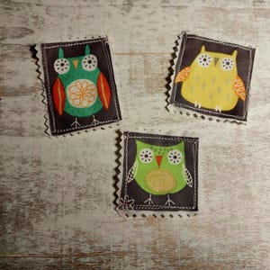 May include: Three fabric owl appliques with black stitching around the edges. The owls are green, yellow, and green with black eyes and white pupils. The owls are on a light wood background.