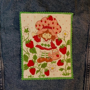 May include: A rectangular fabric patch featuring a cartoon character with brown hair wearing a strawberry-patterned hat and dress, holding a watering can. The patch is surrounded by green strawberry vines and white flowers, finished with a green zigzag border on blue denim.