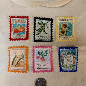 May include: A cream-colored fabric tote bag featuring six rectangular patches with scalloped edges, each depicting vintage-style seed packets for tomato, peas, sunflower, sweetcorn, radish, and sweet peas, with a silver US quarter coin for scale.