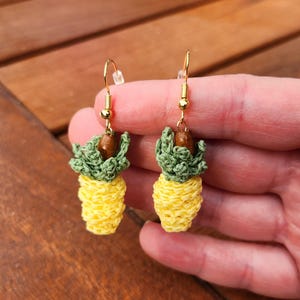 May include: A pair of crocheted pineapple earrings with gold hooks. The pineapples are yellow with green tops and brown centers.