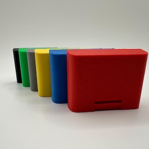 May include: A row of colorful, rectangular, 3D-printed objects. The colors include black, green, gray, yellow, blue, and red. Each object has a textured surface and a small rectangular cutout on the front.