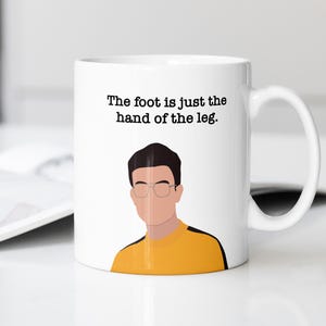 May include: White ceramic mug with a black and white illustration of a man wearing glasses and a yellow shirt. The text "The foot is just the hand of the leg." is printed on the mug.