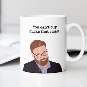 May include: White ceramic mug with a handle, featuring a cartoon illustration of a man with glasses and a beard. The mug has the text "You can't buy ducks that small."