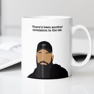 May include: White ceramic mug featuring a graphic of a man with a beard and a black cap with the word "UNREAL". The mug has the text "There's been another revelation in the lab."