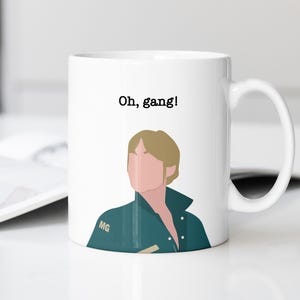 May include: White ceramic mug with a cartoon illustration of a person and the text "Oh, gang!". The person is wearing a teal jacket. The mug has a white handle and is on a white surface.