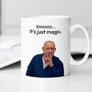 May include: White ceramic mug featuring an image of a man with his finger to his lips, with the text "Shhhhh... It's just magic." printed in black. The mug has a curved handle and is set against a blurred background.