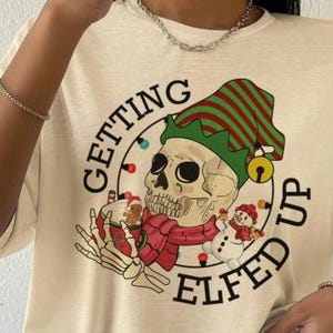 以下が含まれることがあります： ベージュのTシャツに、マグカップを持ったスケルトンエルフのグラフィックプリントが施されています。グラフィックの周りに「Getting Elfed Up」というテキストがプリントされています。