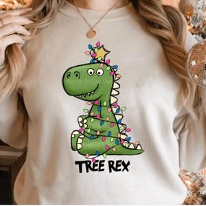 Puede incluir: Una sudadera blanca con un dinosaurio verde que lleva luces de Navidad y el texto "TREE REX".