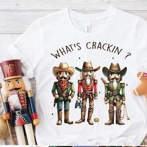 May include: White t-shirt with a graphic of three nutcracker cowboys wearing hats and holding guns. The text "What's Crackin?" is printed above the nutcrackers.
