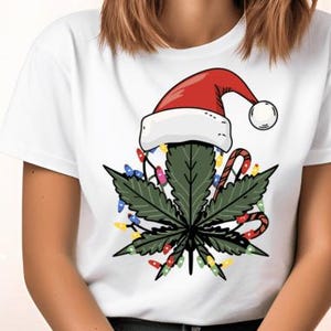 May include: White t-shirt featuring a green marijuana leaf wearing a red Santa hat with white trim. The leaf is adorned with colourful Christmas lights and candy canes.